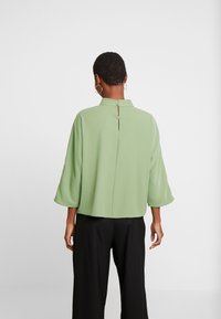 Top verde a maniche corte con colletto alto e chiusura con bottone sul retro. Tessuto strutturato con una superficie liscia, abbinato a pantaloni neri.