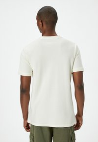 Kortärmad t-shirt med rund halsringning i ljus krämfärg, med en strukturerad tyg och kontrasterande ribbade manschetter. Bakvy visar en avslappnad passform.