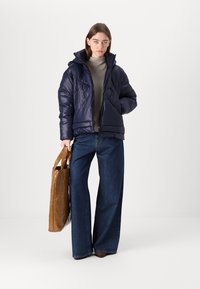 Tumši zils pūkains jaciņa ar spīdīgu apdari, ar rāvējslēdzēju un kapuci. Valka virs pelēka topa un kombinēta ar platu kāju zilo džinsu un brūnu somu.