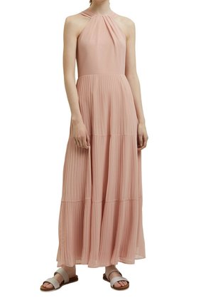 Femme portant une robe maxi rose poudré plissée sans manches avec un col halter et des sandales blanches plates, debout devant un fond blanc.