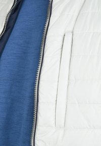 Witte gewatteerde jas met een zilveren rits en een zijzak, gedragen over een blauw structuurshirt.