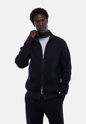 Gilet zippé côtelé de couleur marine en tissu tricoté texturé, avec des manches longues et une fermeture éclair, porté par-dessus une chemise gris clair.