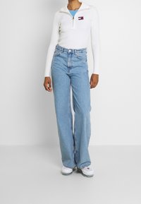 Haut blanc à manches longues côtelé avec une fermeture éclair à l'avant et un logo sur la poitrine, associé à un jean large bleu clair et des baskets blanches.