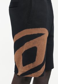 Pantalones cortos negros con un gran diseño abstracto en espiral marrón en el muslo, con bolsillo lateral abierto visible, usados por una persona de piel oscura.