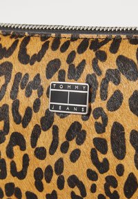 Pochette en simili fourrure à motif léopard avec une texture douce. Elle dispose d'une fermeture éclair noire et d'une étiquette en métal affichant "TOMMY JEANS."