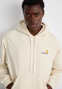 Beige hoodie med snoretræk og frontlomme, der har et lille, farverigt Carhartt-logo på brystet. Blød stoftekstur.