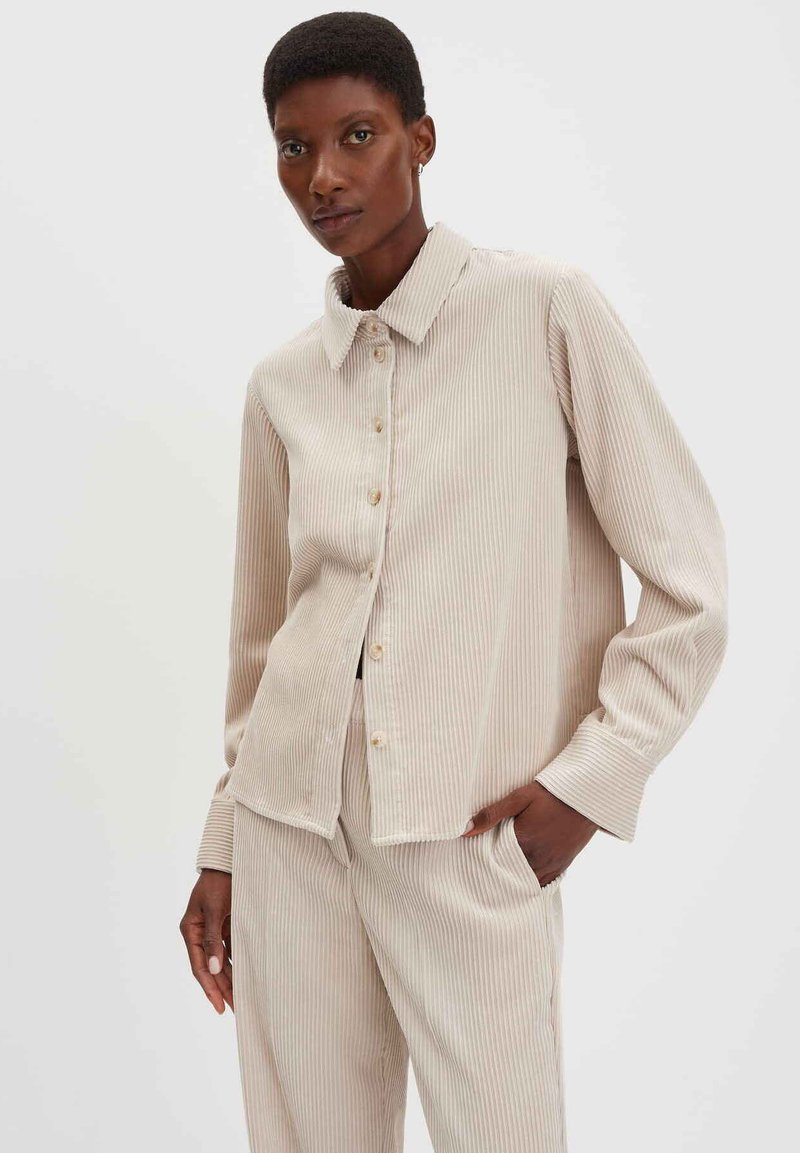 Camicia di velluto a coste beige con colletto, righe verticali, chiusura a bottoni e maniche lunghe, abbinata a pantaloni in velluto a coste della stessa tonalità. Tessuto spesso, vestibilità rilassata.
