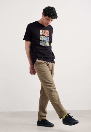 Camiseta negra de manga corta con un diseño abstracto colorido en la parte frontal. Combinada con pantalones marrón claro y zapatos negros con calcetines verdes.