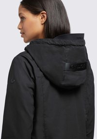 Veste noire à capuche avec une texture lisse et mates. Présente un patch logo à l'arrière et un détail de bouton sur la manche. Design simple.