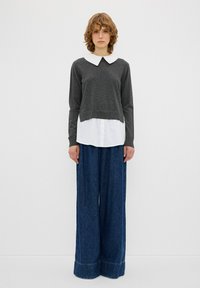Grauer Pullover mit einem weißen Kragen, der über einem weißen Hemd getragen wird, kombiniert mit blauen Jeanshosen im weiten Schnitt und einem umgenähten Saum.