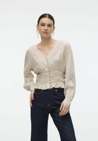 Beige blouse met lange mouwen, een V-hals, een geraffineerde taille, een gerimpelde zoom en knoopdetails, gecombineerd met donkere spijkerbroek.