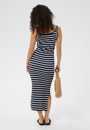 Žena s kovrčavom kosom koja nosi mornarsko plavu i bijelu prugastu haljinu bez rukava, držeći pletenu torbicu, stoji okrenuta leđima prema jednobojnoj pozadini.