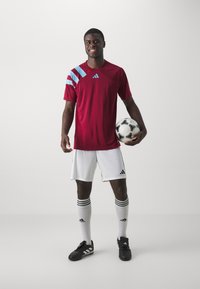 Rotes Fußballtrikot mit kurzen Ärmeln und blauen Akzenten, weiße Shorts, schwarze Fußballschuhe und kniehohe weiße Socken, hält einen schwarz-weißen Fußball.