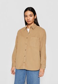 Chemise boutonnée marron clair avec col, poche avant et manches longues. Tissu texturé avec finition naturelle. Modèle la portant.