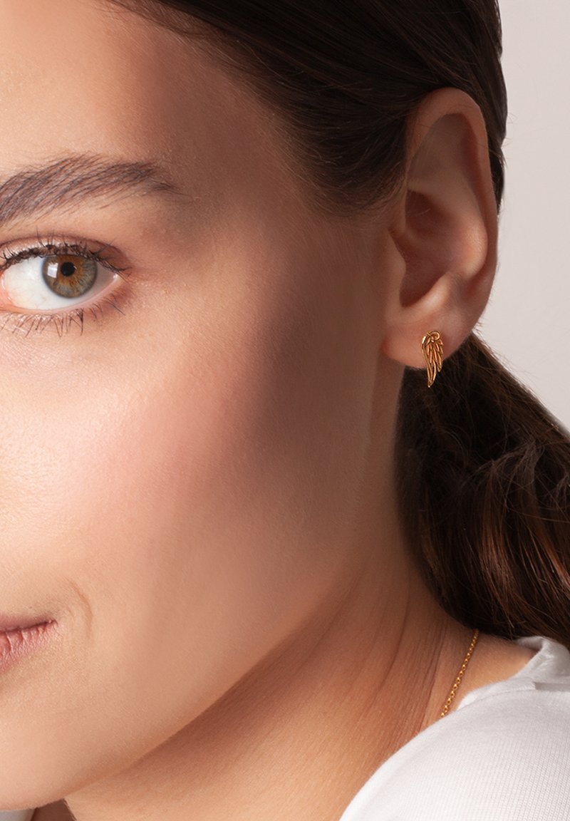 Złoty klips w kształcie skrzydła na uchu modelki, z detalicznymi teksturami przypominającymi pióra i wypolerowanym wykończeniem. Odcień skóry jest neutralny.