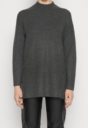 Osoba oblečená v tmavě šedém žebrovaném úpletu s polopriléhavým límcem a dlouhými rukávy, v kombinaci s černými koženými kalhotami.