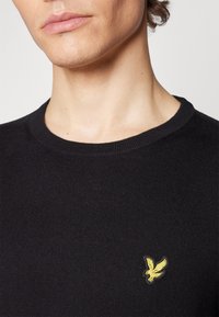 Pull noir en maille avec un col rond et un petit logo d'aigle jaune sur le côté gauche de la poitrine. Texture douce, design ajusté.
