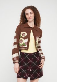 Γυναίκα με καφέ κοντή ζακέτα διακοσμημένη με ηλιοτρόπια και ριγέ μανίκια, κίτρινο τοπ και φούστα με κουμπιά σε μπορντό καρό.