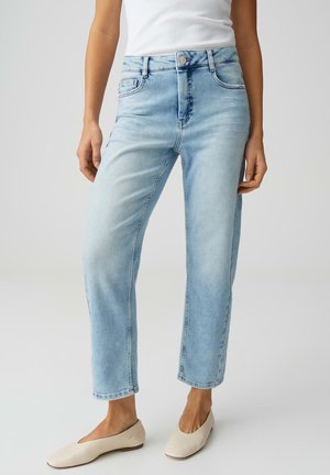 Frau trägt hellblaue, hoch taillierte Jeans mit geradem Bein, weißes ärmelloses Oberteil und beige flache Schuhe, steht vor einem schlichten Hintergrund.