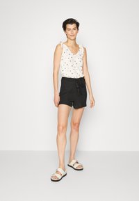Top branco sem mangas com padrão floral e alças atadas, combinado com calções pretos com cinto atado. Footwear é sandálias bege com fivelas.