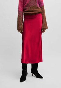 Red satin midi skirt with a smooth texture, featuring a high waist and a straight silhouette. Styled with a brown knit sweater and black boots.