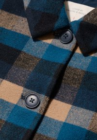 Zažim kuhinjskih gumbov in ovratnika volnenega plašča v modri, bež in črni kariranem vzorcu z vidno nalepko z napisom "Ljubljeno pri mornarjih in umetnikih."