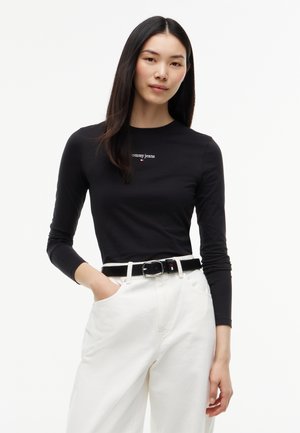 Kobieta w czarnej bluzce z długim rękawem z logo "Tommy Jeans", białe spodnie z wysokim stanem i czarnym paskiem, stoi z jedną ręką w kieszeni.