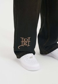 Pantalones de mezclilla negra con un logo bordado en crema en el puño izquierdo, combinados con zapatillas blancas con detalles perforados.