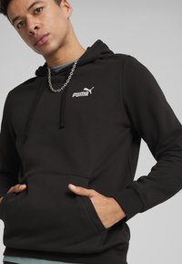 Sweatshirt à capuche noire en tissu doux. Il dispose d'une poche kangourou, de poignets côtelés et d'un logo Puma blanc bien visible sur la poitrine.