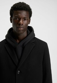 Cappotto in misto lana nero con rever a punta, chiusura a un bottone, indossato sopra una felpa nera con cappuccio, caratterizzato da una texture liscia.