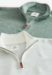 Două pulovere cu fermoar: unul de culoare verde deschis texturat, cu un guler ribbed și un mânere din piele maro, celălalt alb uniform, cu un design similar.