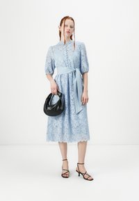 Lyseblå blonde midikjole med pufferm, en knyttet midje-sash og bølget kant. En svart håndveske og svarte sandaler med stropper fullfører looken.