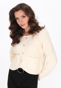 Cardigan crema lavorato a maglia con un motivo testurizzato, dotato di bottoni a toggle in tono oro, due tasche frontali e un orlo arrotondato.