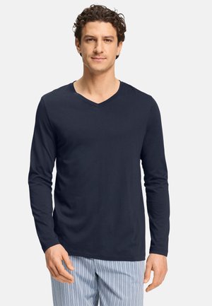Homme portant une chemise bleu marine à manches longues et col en V ainsi qu'un pantalon bleu clair rayé, debout devant un fond uni.