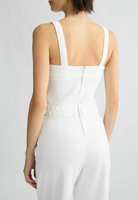 Top sans manches blanc avec une silhouette ajustée, présentant une fermeture éclair au dos et un détail tressé au niveau de la taille. Tissu de texture lisse.