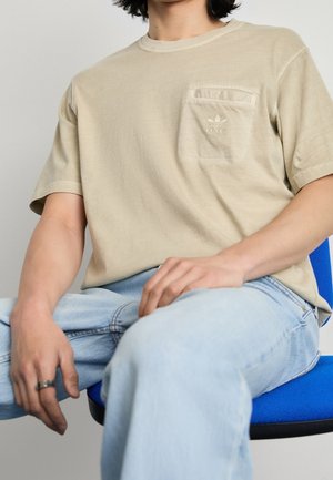 Beżowa bawełniana koszulka z okrągłym dekoltem, krótkim rękawem i kieszonką na piersi. Posiada subtelne logo "adidas" na kieszeni.