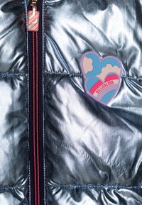 Section de blouson matelassé en métal argenté avec une fermeture éclair rose contrastante et un patch en forme de cœur avec des nuages et le texte "BILLIEBLUSH".