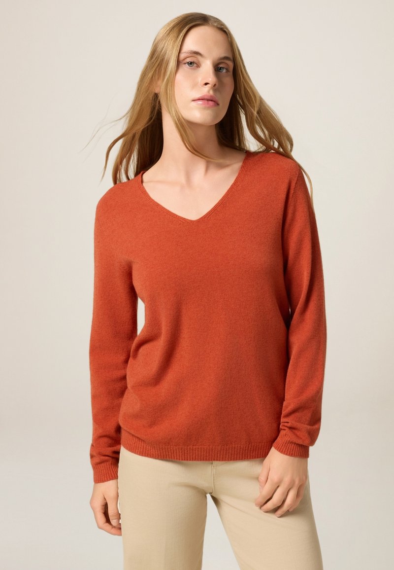Pull à col en V, à manches longues, en tissu orange doux et texturé. Présente des poignets ajustés et une coupe décontractée, assorti à un pantalon de couleur claire.