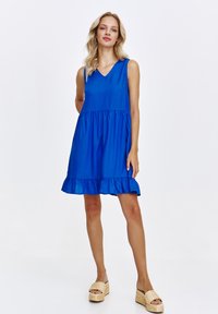 Niebieska sukienka bez rękawów z dekoltem w kształcie litery V i rozszerzanym dołem z falbankami. Gładka tekstura, zwiewny krój, długość do kolan.