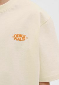 Krátký rukáv béžové tričko vyrobené z bavlny. Na hrudi je vyšívaný nápis v oranžové barvě "ORIGINALS 1990". Jemná textura, volný střih.