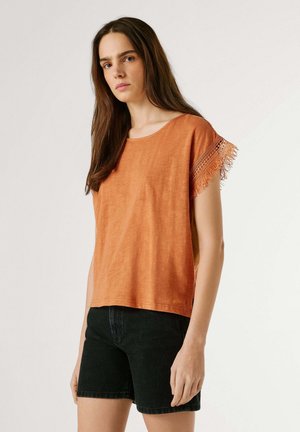 Pitkähiuksinen nainen, jolla on päällään oranssi lyhythihainen paita, jossa hapsuista yksityiskohtaa, ja mustat shortsit, seisoo yksinkertaisen taustan edessä.