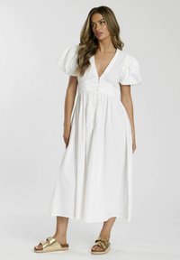 Weißes Baumwollkleid mit tiefem V-Ausschnitt, Puffärmeln und Knopfdetails. Tragen Sie es mit goldenen Sandalen und es hat einen fließenden, knöchellangen Rock.