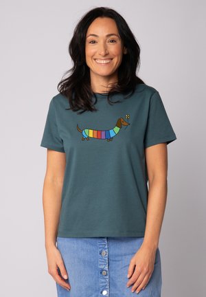 Lächelnde Frau trägt ein türkisfarbenes T-Shirt mit einem Dackel mit Regenbogenstreifen, der eine gelbe Blume hält, kombiniert mit einem blauen Jeansrock mit Knöpfen.