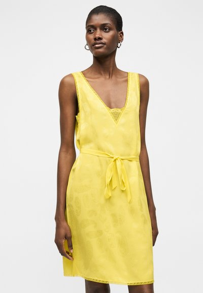 Γυναίκα με κίτρινο αμάνικο φόρεμα με V-λαιμόκοψη, ζώνη στη μέση και απαλό μοτίβο πάισλεϊ, στέκεται μπροστά σε απλό λευκό φόντο.