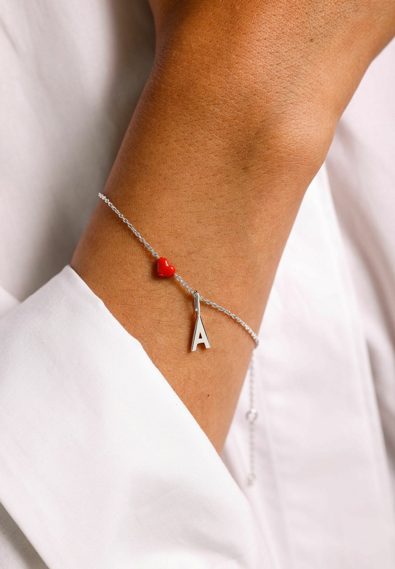 Silverkedja armband med ett rött hjärta charm och en silverbokstav "A" hängande, mot hudton, med en slät, polerad finish.