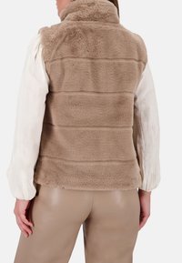 Chaleco de piel sintética beige con cuello alto, costuras horizontales y textura suave, combinado con una blusa de color claro y pantalones ajustados.