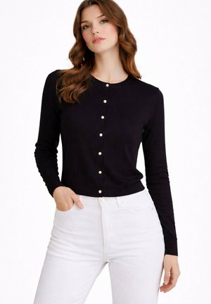 Femme aux cheveux bruns ondulés portant un haut noir à manches longues boutonné et un pantalon blanc taille haute, debout avec une main dans la poche.