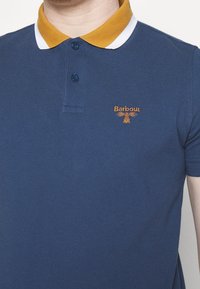 Homme portant un polo bleu marine avec un col jaune moutarde et blanc, avec un petit logo Barbour brodé sur la poitrine.