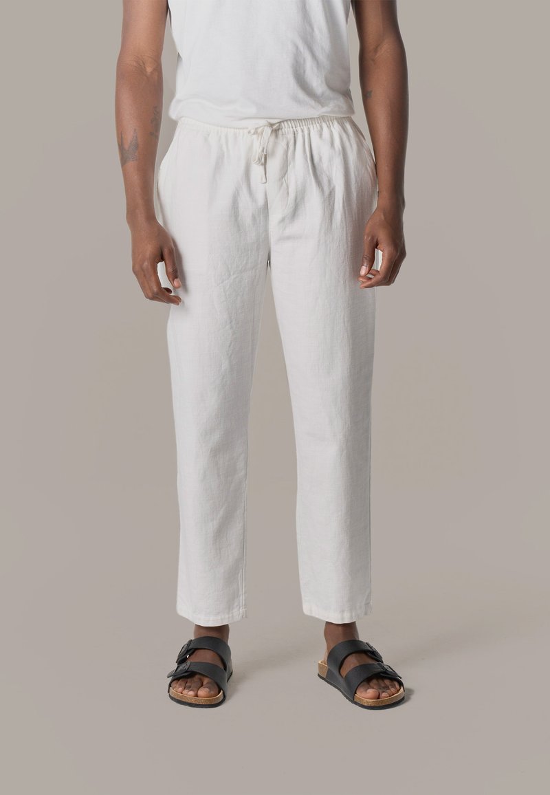 Persona con pantalones blancos con cordón y sandalias negras de doble correa, de pie contra un fondo neutro y simple.