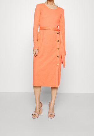 Femme portant une robe midi corail à manches longues, boutons sur le côté, taille nouée, et sandales à talons blocs nude, debout devant un fond blanc.